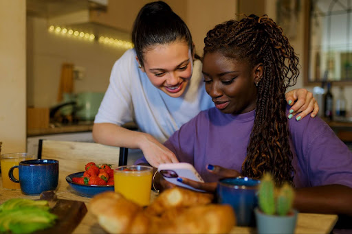 Mulher branca apoiada em mulher negra que segura um celular sentada a mesa